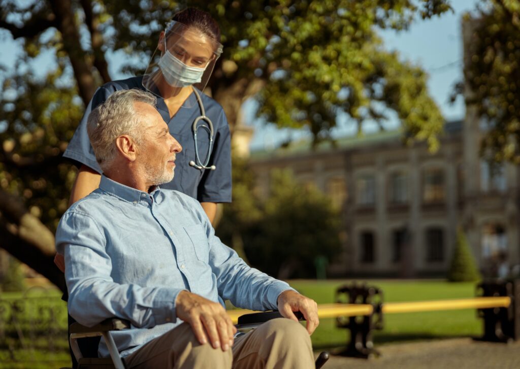 Caring young nurse wearing face shield and mask talking to mature man, pushing patient in wheelchair