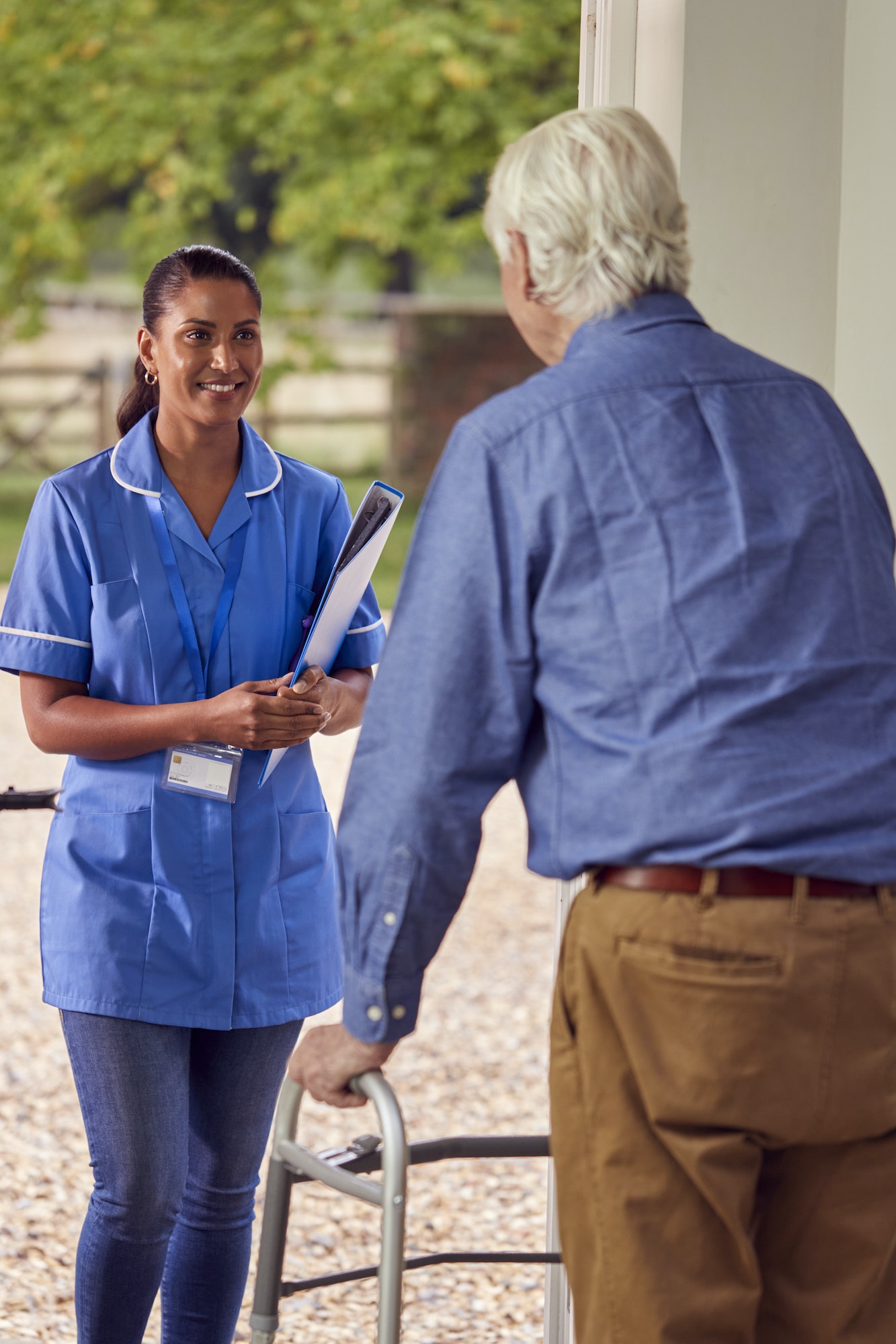 Senior Man At Home Using Walking Frame Greeting Female Nurse Or Care Worker In Uniform At Door